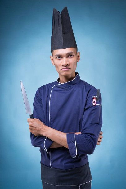 Портрет азиатского шеф-повара в синей форме, черном фартуке и черной шляпе на синем фоне
 - Фото, изображение