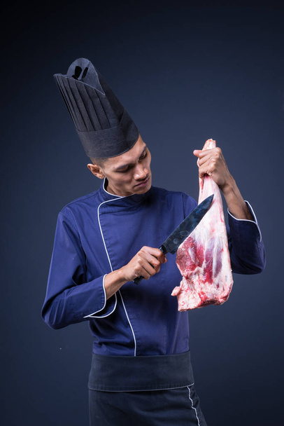 Портрет азіатського виконавчого шеф-кухаря в його синій формі, чорний фартух і чорний капелюх на сірому тлі з ягнячою ногою. - Фото, зображення