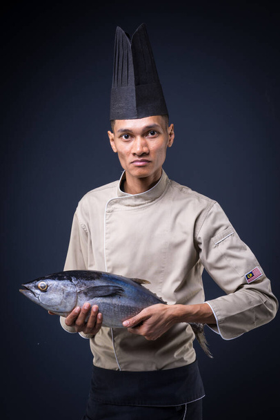 Egy ázsiai főszakács portréja világosbarna egyenruhában, fekete kötényben és fekete kalapban szürke háttéren, csíkos tonhallal. - Fotó, kép