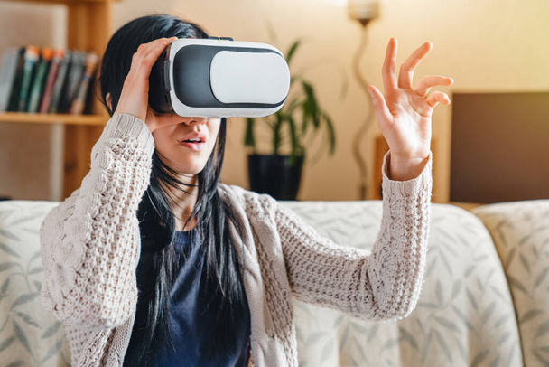 Frau berührt etwas mit Virtual-Reality-Brille und sitzt zu Hause auf der Couch - Foto, Bild