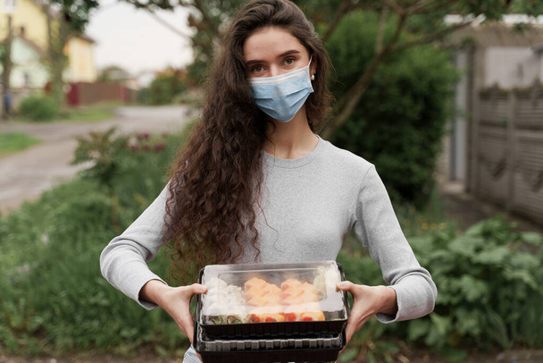 Girl courier in medical mask with 2 sushi boxes stands in front of car. Sushi set in box healthy food delivery service by car. Japanese cuisine: rolls, soy sauce, wasabi. 1+1 advert for restaurant - Photo, Image