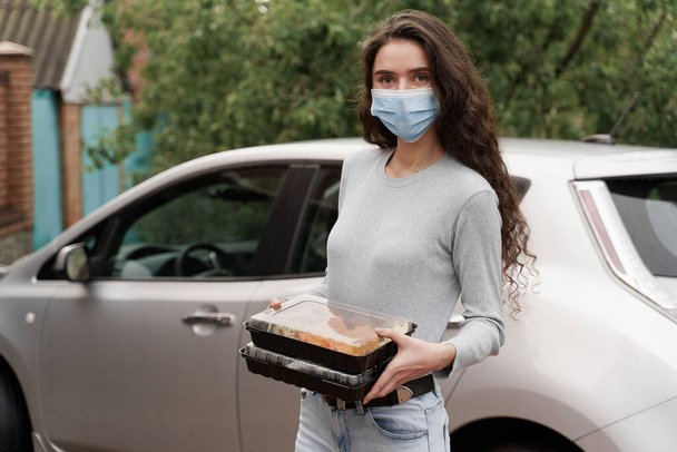 車での宅配サービスも充実しています。車の前には2つの寿司箱が付いた医療用マスクの女の子の宅配便が立っています。日本料理：巻き、醤油、わさび。持ち帰り用の1 + 1広告 - 写真・画像