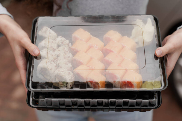 Κοντινό σούσι σε κουτί υγιεινά τρόφιμα παράδοση σε απευθείας σύνδεση υπηρεσία. Το κορίτσι κρατάει 2 σούσι στα χέρια του. Ιαπωνική κουζίνα: ρολά, σάλτσα σόγιας, wasabi. 1 + 1 αγγελία για εστιατόριο - Φωτογραφία, εικόνα