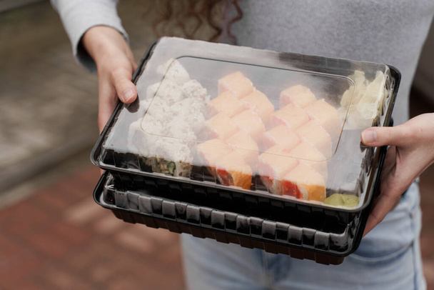 Κοντινό σούσι σε κουτί υγιεινά τρόφιμα παράδοση σε απευθείας σύνδεση υπηρεσία. Το κορίτσι κρατάει 2 σούσι στα χέρια του. Ιαπωνική κουζίνα: ρολά, σάλτσα σόγιας, wasabi. 1 + 1 αγγελία για εστιατόριο - Φωτογραφία, εικόνα