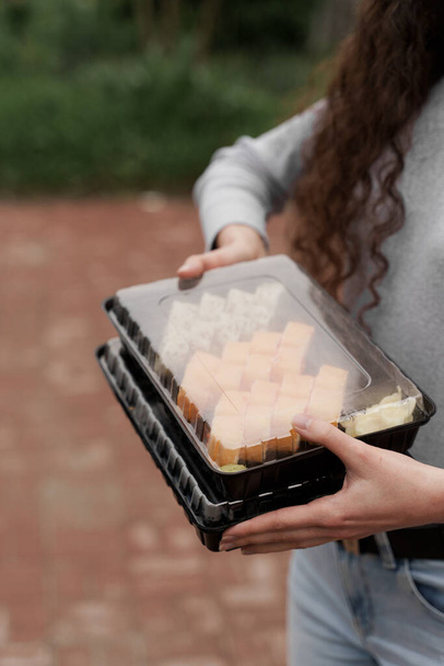 Zbliżenie sushi w pudełku zdrowej żywności dostawy usług online. Dziewczyna posiada 2 zestawy sushi w rękach. Kuchnia japońska: bułki, sos sojowy, wasabi. 1 + 1 reklama restauracji. Zdjęcie pionowe dla sieci społecznościowych - Zdjęcie, obraz