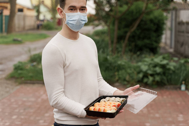 Sushi set in doos gezonde voeding levering online service met de auto. Man koerier met medisch masker opent sushi doos. Japanse keuken: broodjes, sojasaus, wasabi. 1 + 1 advertentie voor afhaalmaaltijden - Foto, afbeelding