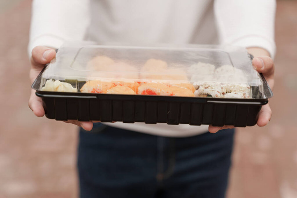 Closeup sushi in box healthy food delivery online service. Man in white clothes gives sushi set to you. Japanese cuisine: rolls, soy sauce, wasabi. Advert for restaurant - Photo, Image