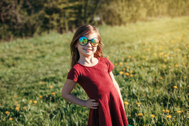 Χαρούμενη χαμογελαστή λευκή κοπέλα με αστεία γυαλιά ηλίου που ποζάρει σε εξωτερικό χώρο. Χαριτωμένο αξιολάτρευτο παιδί που διασκεδάζει έξω. Χαρούμενη παιδική ηλικία. Ειλικρινής θετική συγκίνηση.  - Φωτογραφία, εικόνα