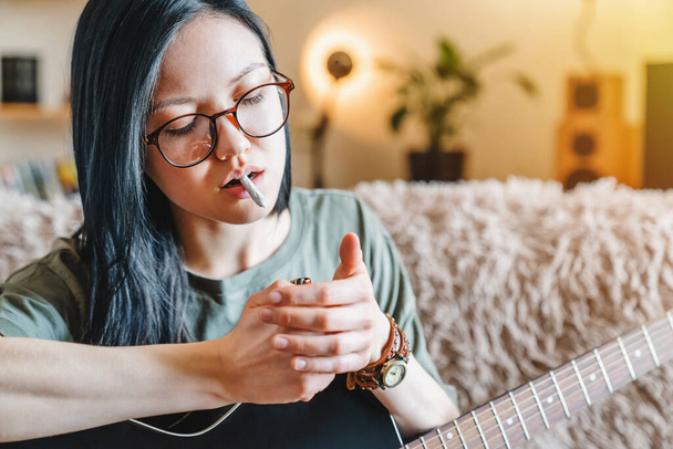 Close up of young girl lighting marijuana joint while holding guitar at home - Foto, Imagen