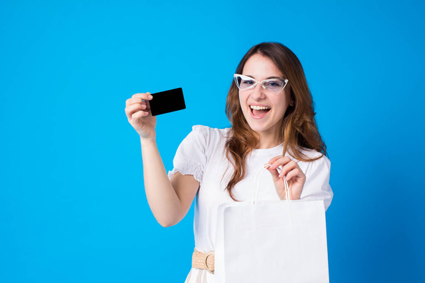 Portrét šťastné mladé brunetky ženy zuby s úsměvem ve slunečních brýlích a letní oblečení s černou bankovní kreditní kartou a bílou prázdnou nákupní taškou, při pohledu do kamery, izolované na modrém pozadí.Nakupování - Fotografie, Obrázek