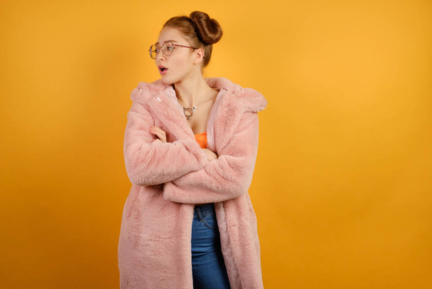 A red-haired girl in an orange top, round glasses and a pink fur coat looks away, crossing arms over chest - Fotografie, Obrázek