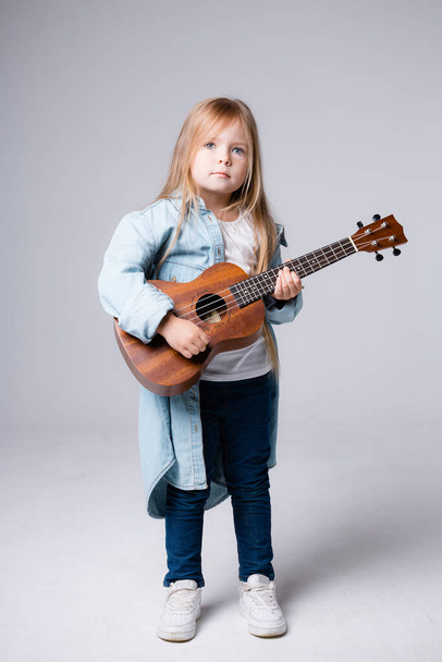 Dívka dítě britského vzhledu s dlouhými vlasy stojí v modré košili s ukuleles v rukou na šedém pozadí - Fotografie, Obrázek