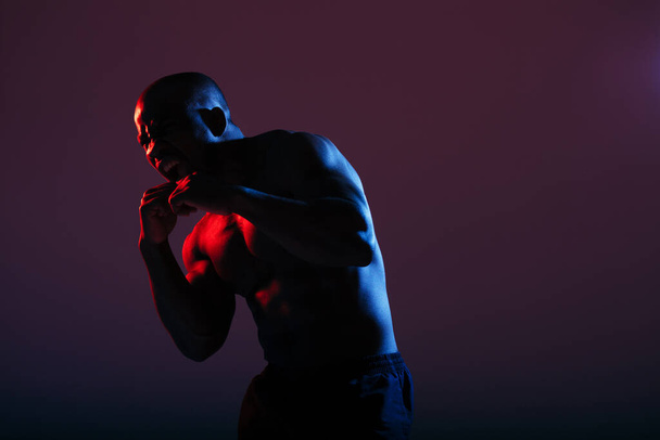 An African American in a red-blue neon color with a naked torso stood in a combat pose in the studio. Advertising sports shooting - Φωτογραφία, εικόνα