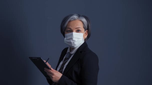 Businesswoman with grey hair in protective mask holding and looking at tablet standing in business suit sideways looking at camera. Health care concept. Business concept. Prores 422 - Séquence, vidéo