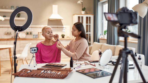 Miracles of beauty. Asian female blogger applying makeup on middle aged woman face using cosmetic products on the table. Two women recording a tutorial video for beauty blog - Photo, image