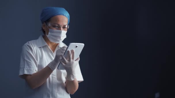 The doctor in a protective mask goggles and a uniform holds in his hands. Tablet editing information of the patient introducing new data. Health concept. Prores 422 - Séquence, vidéo
