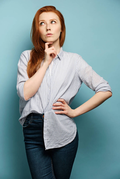 A red-haired girl stands on a blue background in a striped shirt and looks thoughtfully to the side, putting her chin on finger - Фото, зображення