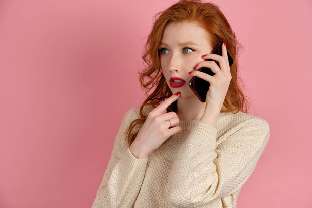 Red-haired girl stands on a pink background in a light sweater with red lips and speaks on the phone, raising a finger to her lips - Photo, Image