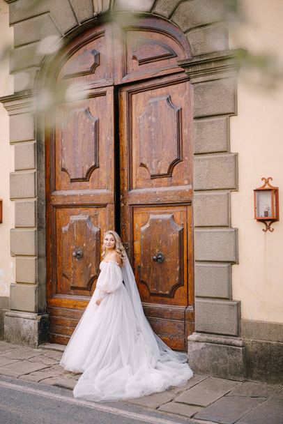 Piękna panna młoda w dużej białej sukni ślubnej i długim welonie stoi przed ogromnymi drewnianymi, zabytkowymi drzwiami starej włoskiej Złoczyńcy Toskanii, Florencji. - Zdjęcie, obraz