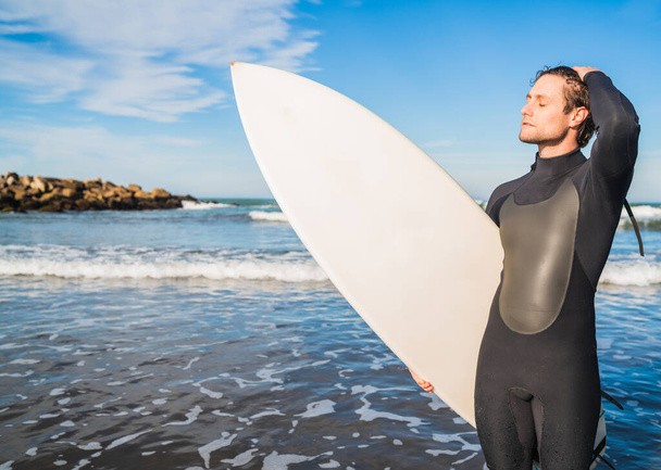 Young surfer standing in the ocean with his surfboard in a black surfing suit. Sport and water sport concept. - Foto, Bild