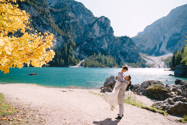 Menyasszony és vőlegény egy őszi fa alatt, tüzes sárga lombozattal, az olaszországi Lago di Braies-ben. Úticél esküvő Európában, a Braies tónál. Az ifjú szerelmesek táncolnak és pörögnek.. - Fotó, kép
