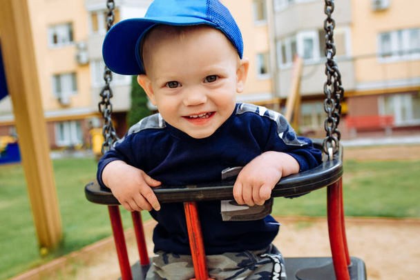 Lustiger und gutaussehender Junge freut sich, fährt auf Kinderschaukel, spielt auf dem Spielplatz - Foto, Bild