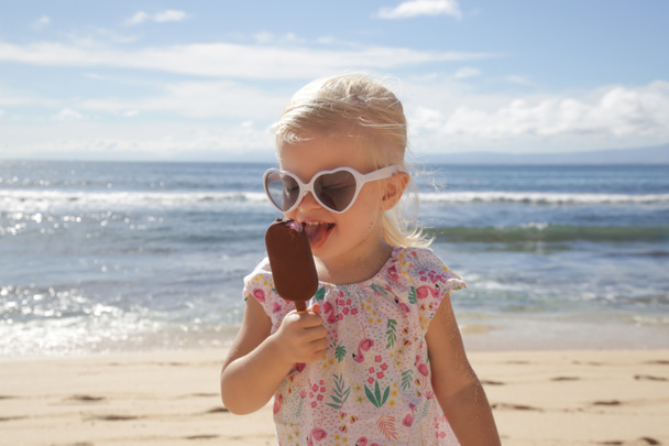 Urocza dziewczynka jedząca lody. Portret dziecka noszącego okulary przeciwsłoneczne i trzymającego lody z pięknym błękitnym morzem i niebem za plecami. Wesołych wakacji na plaży. - Zdjęcie, obraz