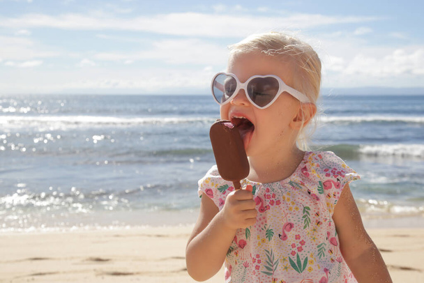 Adorable niña pequeña comiendo helado. Retrato de un niño con gafas de sol y sosteniendo un helado con hermoso mar azul y cielo detrás. Feliz vacaciones de verano en la playa
. - Foto, Imagen