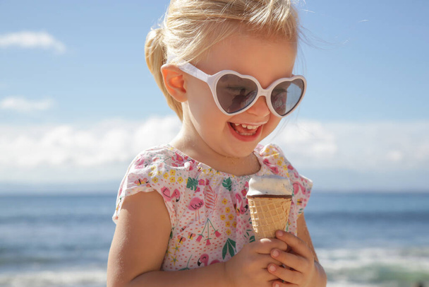 Imádnivaló kislány jégkrémet eszik. Napszemüveges gyermek portréja, kezében egy jégkrémmel, gyönyörű kék tengerrel és égbolttal. Boldog nyári vakációt a strandon. - Fotó, kép