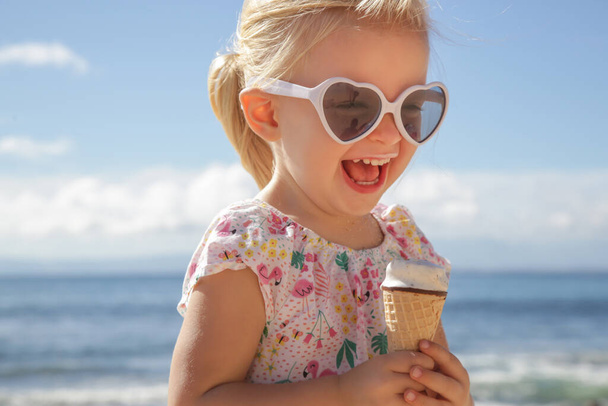 Adorable toddler girl eating ice cream. Portrait of child wearing sunglasses and holding an ice cream with beautiful blue sea and sky behind. Happy summer vacation on the beach. - Foto, Imagen