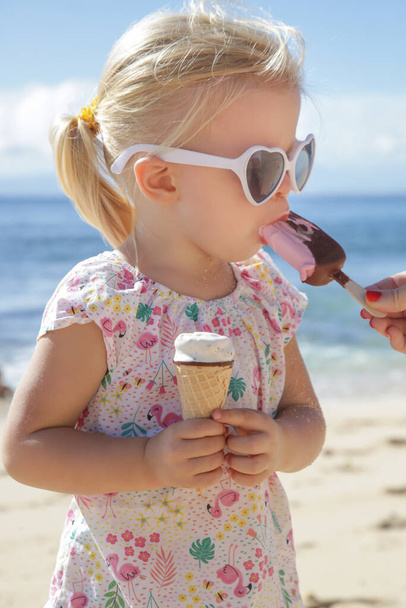 愛らしい幼児の女の子はアイスクリームを食べています。サングラスをかけ、美しい青い海と空の後ろにアイスクリームを持っている子供の肖像画。ビーチでの幸せな夏の休暇. - 写真・画像