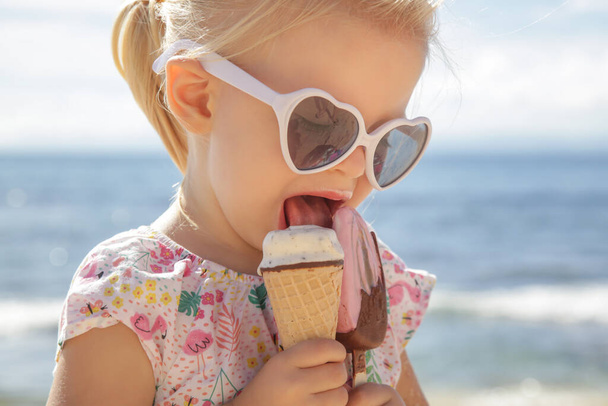 Adorable toddler girl eating ice cream. Portrait of child wearing sunglasses and holding an ice cream with beautiful blue sea and sky behind. Happy summer vacation on the beach. - Φωτογραφία, εικόνα