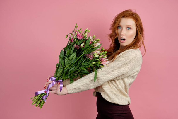 A red-haired girl is standing on a pink background, holding a large bouquet and opening mouth in surprise, looking at the camera - Foto, afbeelding