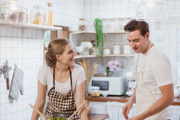 Happy & smiling attractive young cute caucasian couple in love enjoying cooking healthy salad in kitchen at home together. Happiness family of beautiful romantic married lover or marriage relationship - Foto, Imagen