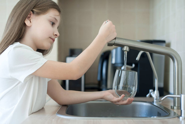 Child is holding a glass under stream of clear transparent cold water from a tap. Close up shot of a young girl pouring a glass of fresh water from a kitchen faucet. Healthy nutrition. World Water Day - Zdjęcie, obraz