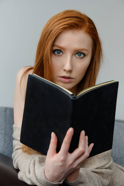 The red-haired girl sits leaning on the arm of the sofa and looks over the open black book in her hands. - 写真・画像