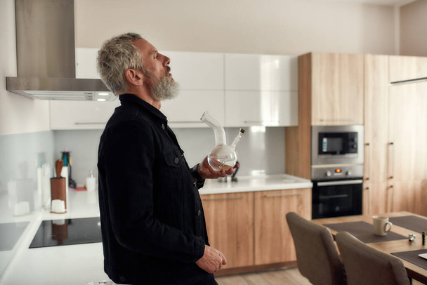 Feel everything. Bearded midle-aged man holding a bong or glass water pipe while smoking marijuana, standing in the kitchen. Cannabis and weed legalization concept - Photo, Image