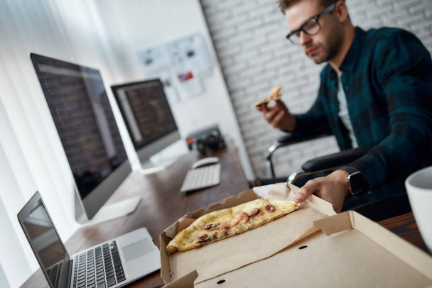 Time for a break. Young male web developer in a wheelchair eating pizza while sitting at his workplace in the modern office. Disabled male programmer having a lunch - Foto, Imagem