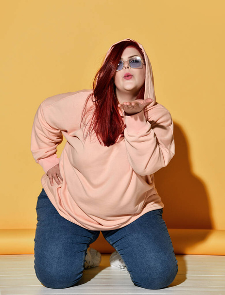 Junge rothaarige übergewichtige Frau in stylischem Kapuzenpulli und Jeans posiert auf dem Boden und schickt uns einen Kuss - Foto, Bild