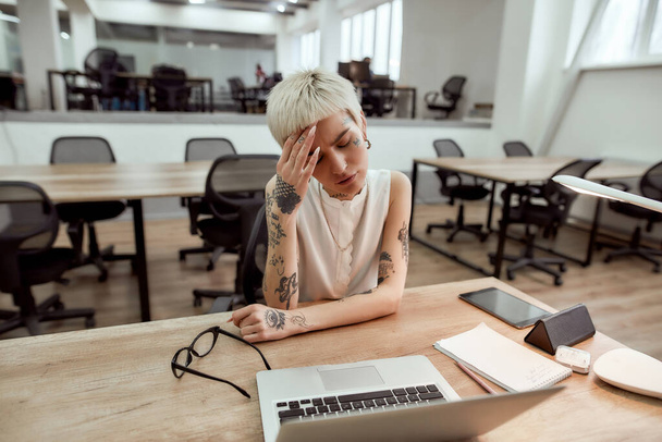Stressful working day. Young tired tattooed business woman keeping eyes closed, touching her head, suffering from the headache while sitting at her working place in the modern office - Foto, Imagem