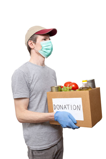 volunteer in medicine mask, protective gloves holding grocery food in carton donation box: vegetable oil, grape, tin cans, tomatoes. Coronavirus relief funds, charity help, delivery, local support - Photo, Image