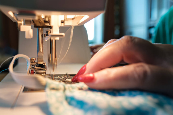 Las manos femeninas cosen una máscara protectora utilizando una moderna máquina de coser eléctrica. Concepto de seguridad durante una epidemia viral
. - Foto, Imagen