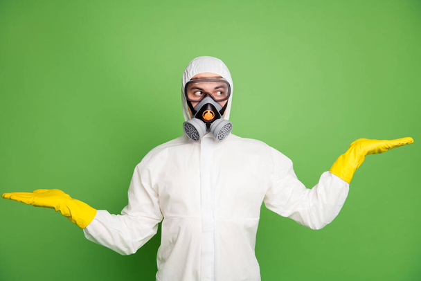 Portrait of uncertain man hold hand look copyspace demonstrate ncov ads promotion wear white latex hazmat uniform isolated over green color background - Zdjęcie, obraz