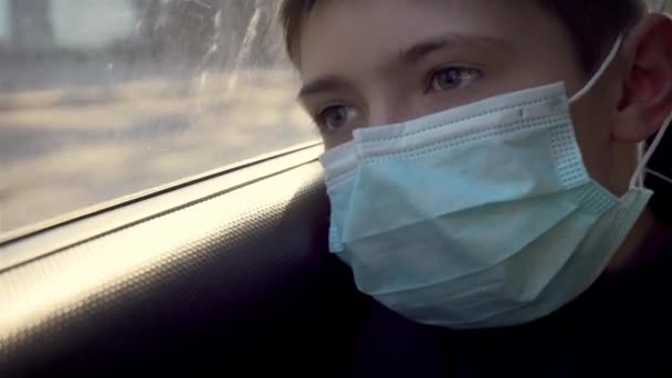 Portrait young boy in medical mask looks out moving car window on street. Self-isolation in quarantine - Materiał filmowy, wideo