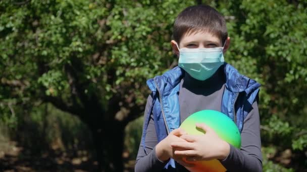 verveeld kind jongen in beschermende masker op gezicht thuis op achtertuin op quarantaine met gekleurde bal - Video