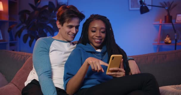 Young couple looking at smartphone and using funny apps while spending time together on sofa. Smiling guy and girl relaxing and scrolling social media news feed . Concept of leisure. - Video