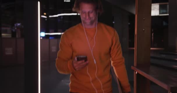 Caucasian man wearing casual clothes and earphones, out and about in the city streets during a night, using his smartphone and holding a skateboard. - Filmmaterial, Video