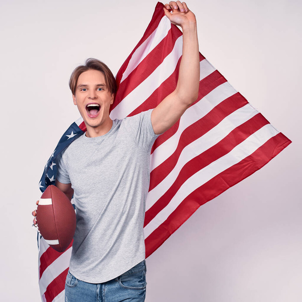 Image of a young handsome guy, in casual clothes, holds the American flag and ball for American football, runs and is smiling, screaming with joy. Emigrant concept, American symbolism, July 4th. - Fotoğraf, Görsel