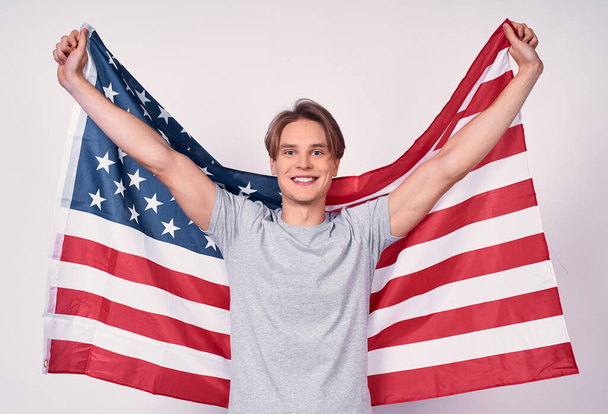 A handsome young slender guy, in casual clothes, is holding an American flag, looking at the camera and smiling, posing on a white background. - 写真・画像