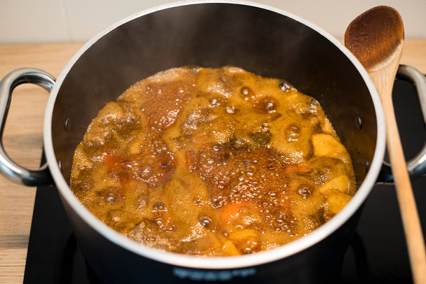 Apricot jam preparation in a black pot. Fruit preserve boiling in a big casserole with a wooden spoon. - Photo, Image